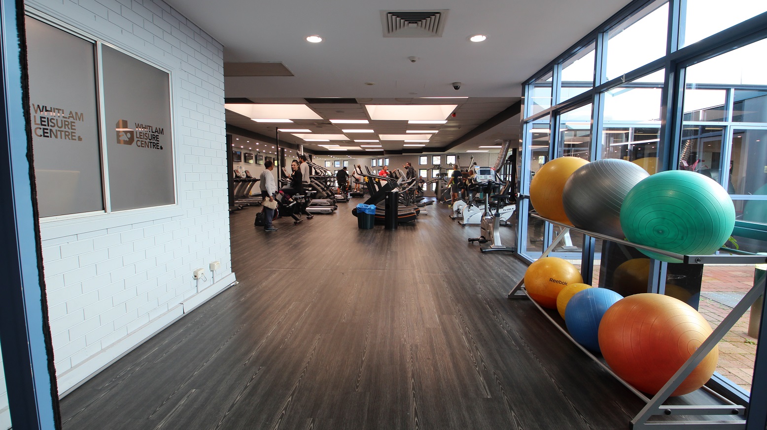 Left of the narrow corridor leading to the fitness facility is made of a white brick wall, while on the opposite side are glass walls and gym balls arranged in two layers of metal platforms. The first machines that guests could see were treadmills. The room is well-lit by square lights at the ceilings.