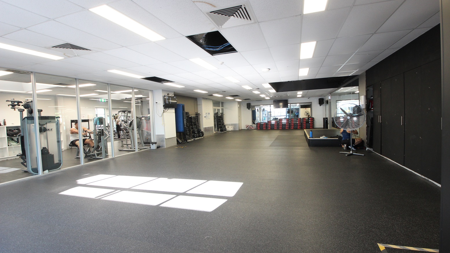 The weight training area, a group exercise studio, and a spin studio at Whitlam Leisure Centre. A large, black rectangular block is placed in the center right after the stand fan. Weight-lifting equipment is arranged against the walls