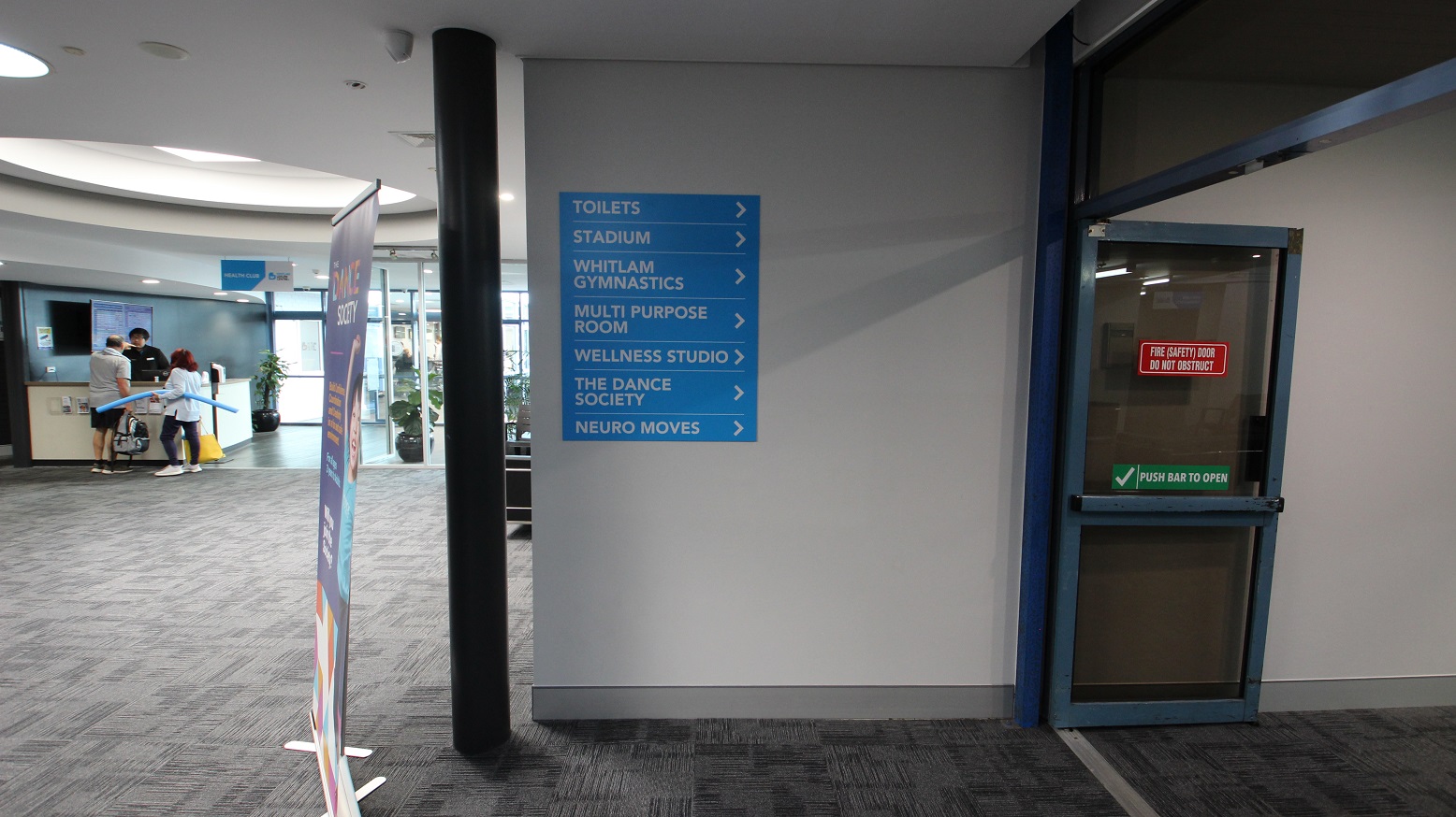 Open, sliding, fire double doors. On the right of these doors is a wall with a class schedule for Bruins Liverpool Skills and Development basketball training and black chairs. There are also a number of chairs settled against the walls at the end of the corridor.