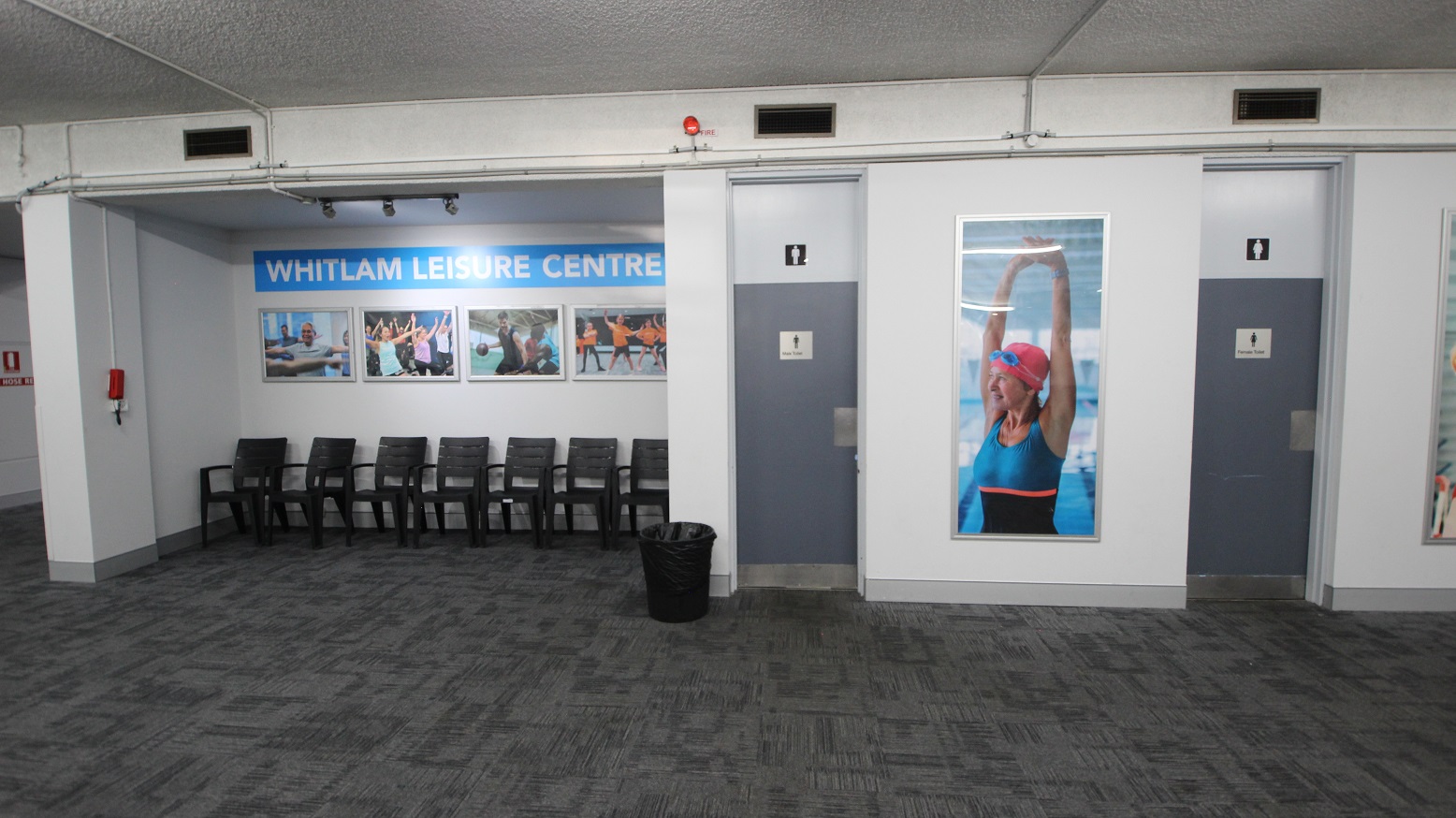 In between the separate toilet rooms for males and females is a photograph of a woman wearing swimming goggles and a swimming cap. Outside the male toilet room is a small rubbish bin. On the left side of the toilet section are black chairs for guests to sit on while waiting for their turn.