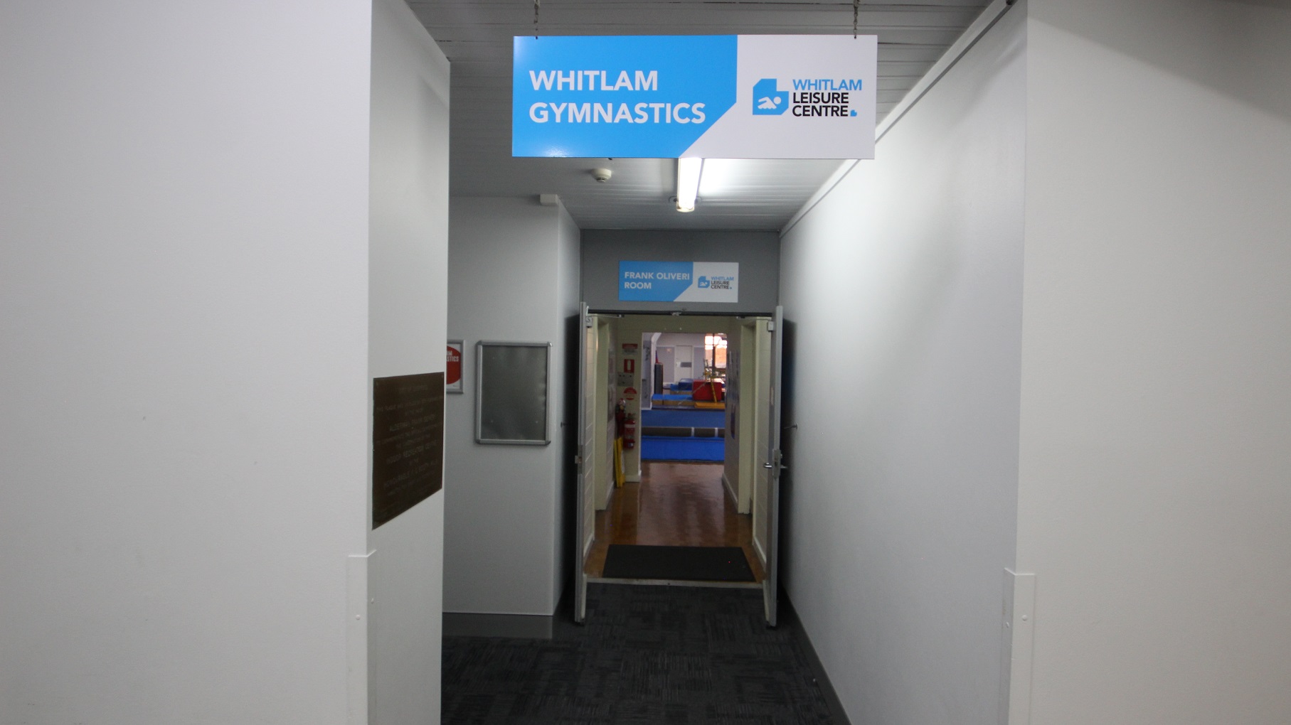 Corridor leading towards Whitlam Gymnastics. The walls are clean and are painted by white. Above the first open door is the signboard containing Frank Oliveri Room, and on the next door is a fire distinguisher mounted on the lower part of the wall. There are large blue mats inside the Gymnasium.