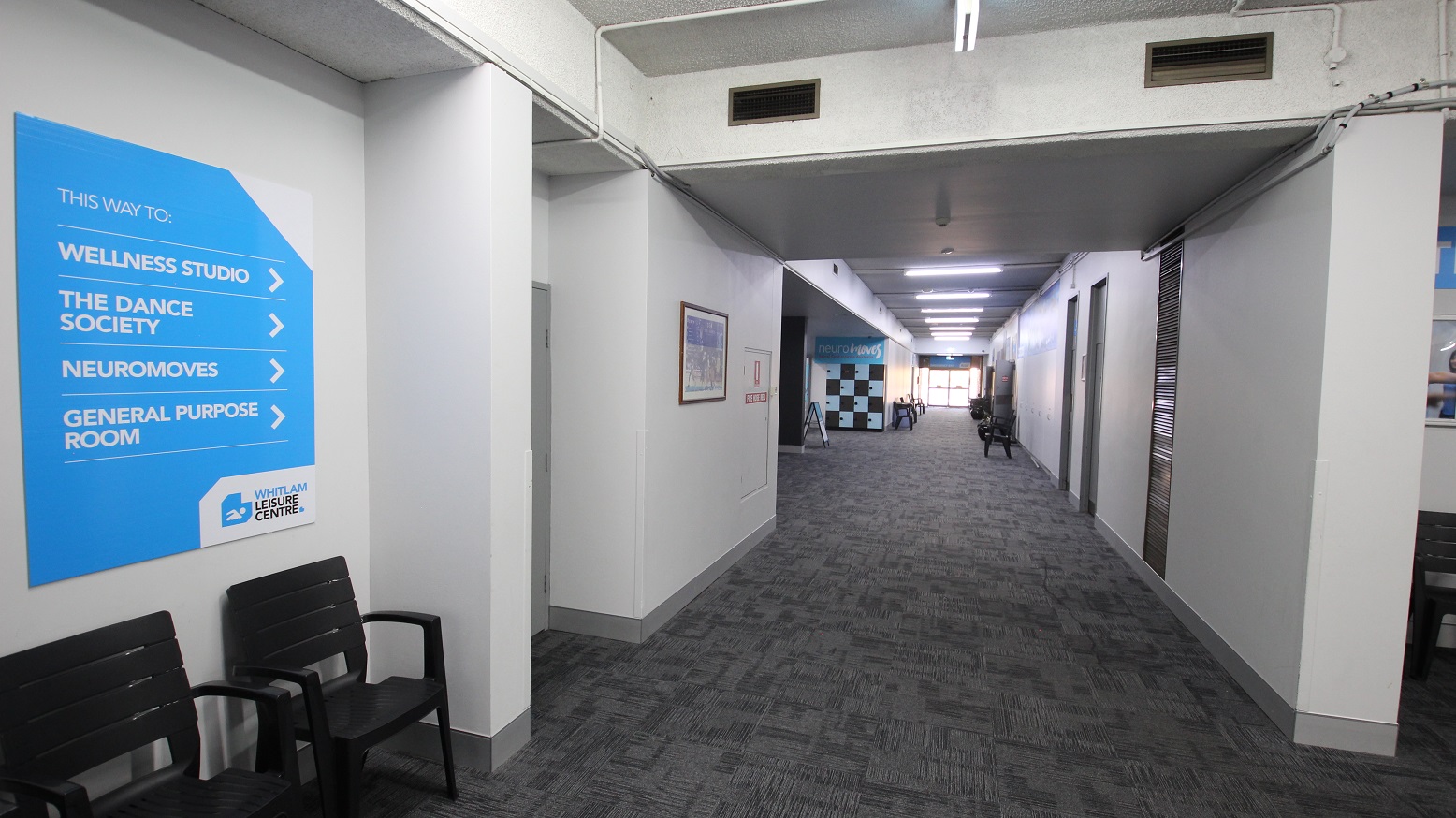 Corridor leading to the Wellness studio. Similar to the other floors of the different kinds of amenities inside the Whitlam Leisure Centre, this also has a grey tiled floor with zigzag lines as a design. A number of black chairs and locker cabinets are placed on the sides of this corridor.