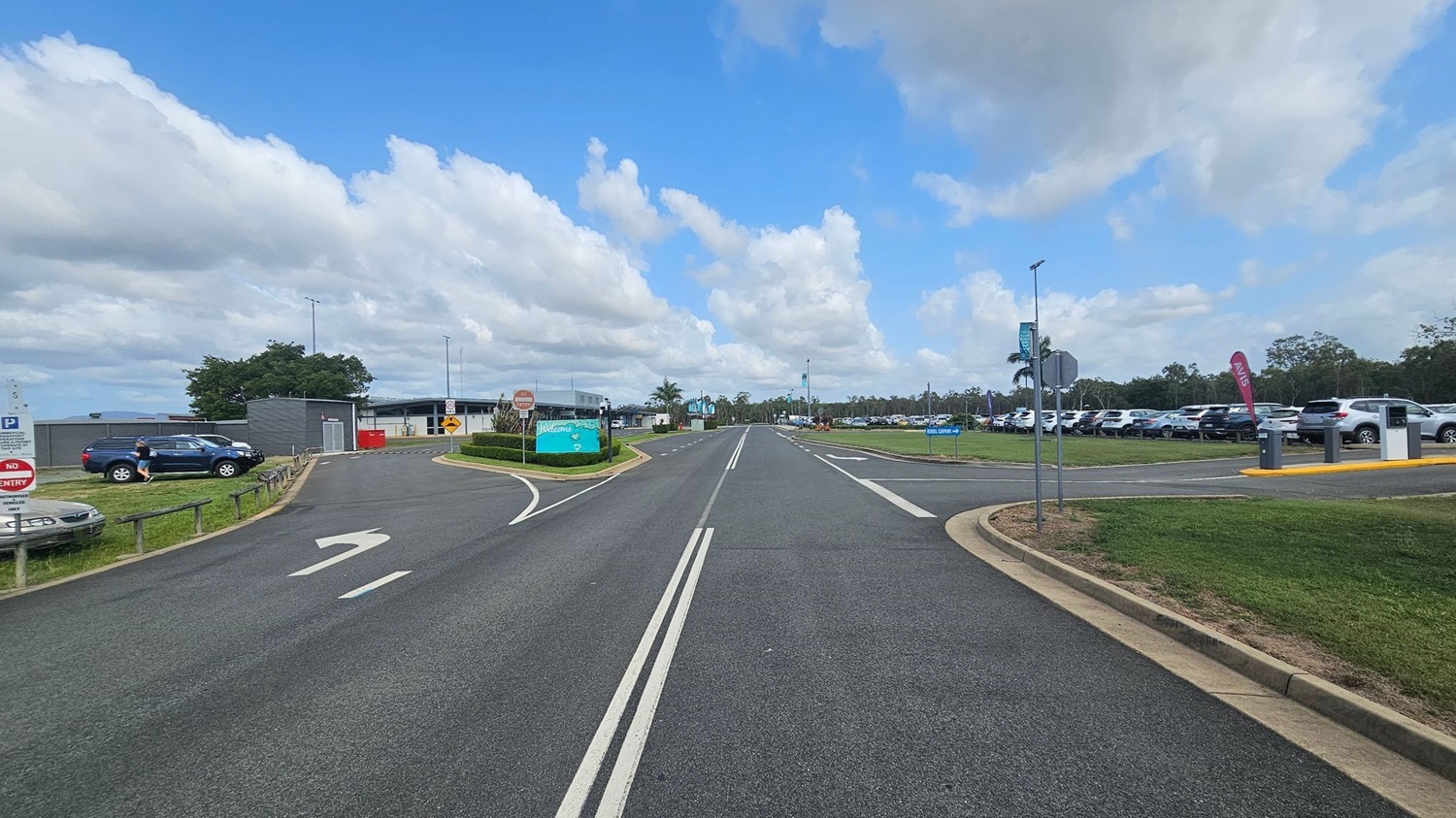 A road with double white lines runs through the centre, flanked by grassy areas. On the left, a car park with a few vehicles and a "No Entry" sign. A person walks near a dark SUV. A building with a grey facade is visible further back. On the right, a larger car park filled with vehicles, a sign, and a flag. Trees and a palm tree are in the background. The sky is partly cloudy.