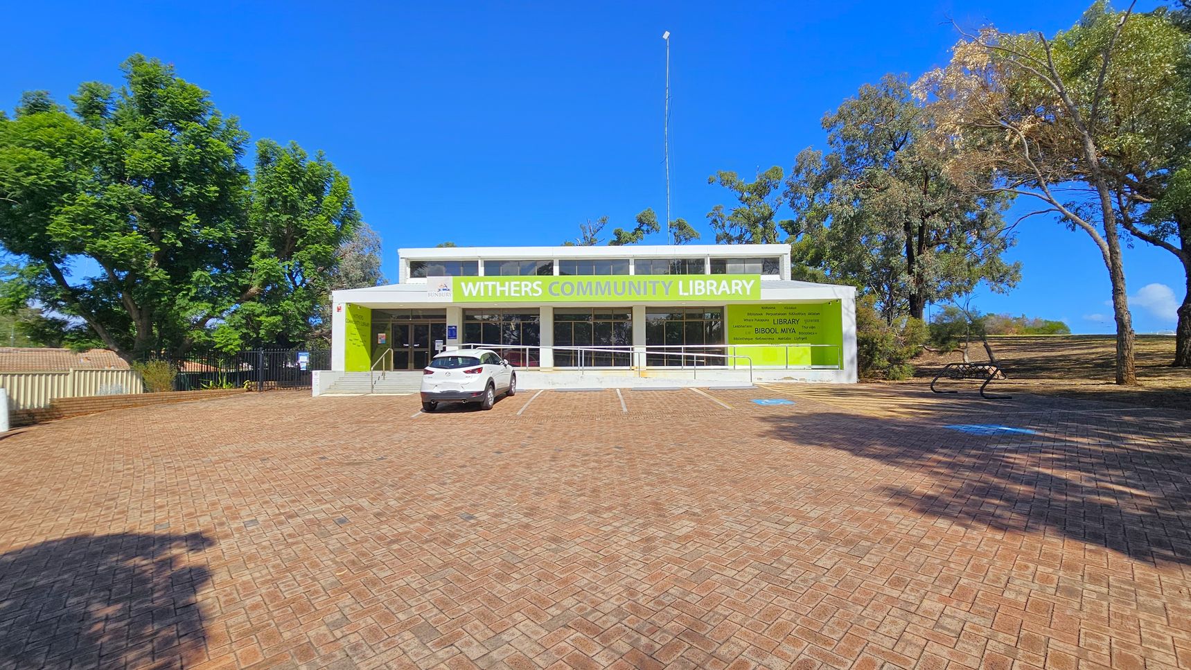 A single-storey building with a sign reading "Withers Community Library" in green and white. The facade is modern with large windows. A white car is parked in front on a brick-paved area. On the left, there are large green trees, and on the right, more trees and a bench. The ground is paved with bricks, and the sky is clear and blue.