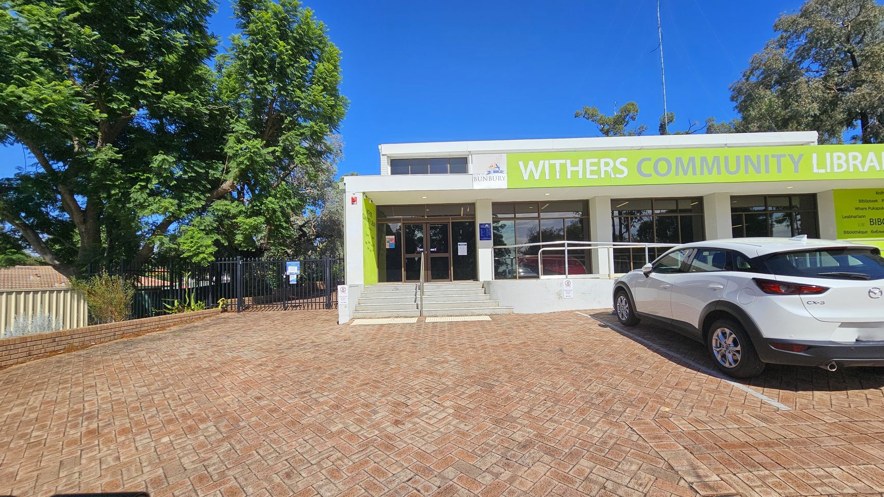 A building with a sign reading "Withers Community Library" in large green letters is in the centre. The entrance features glass doors and a set of steps leading up to it. On the right, a white car is parked on a brick-paved area. To the left, there are large green trees and a black metal fence. The building has a flat roof and large windows. The foreground consists of a brick-paved surface.
