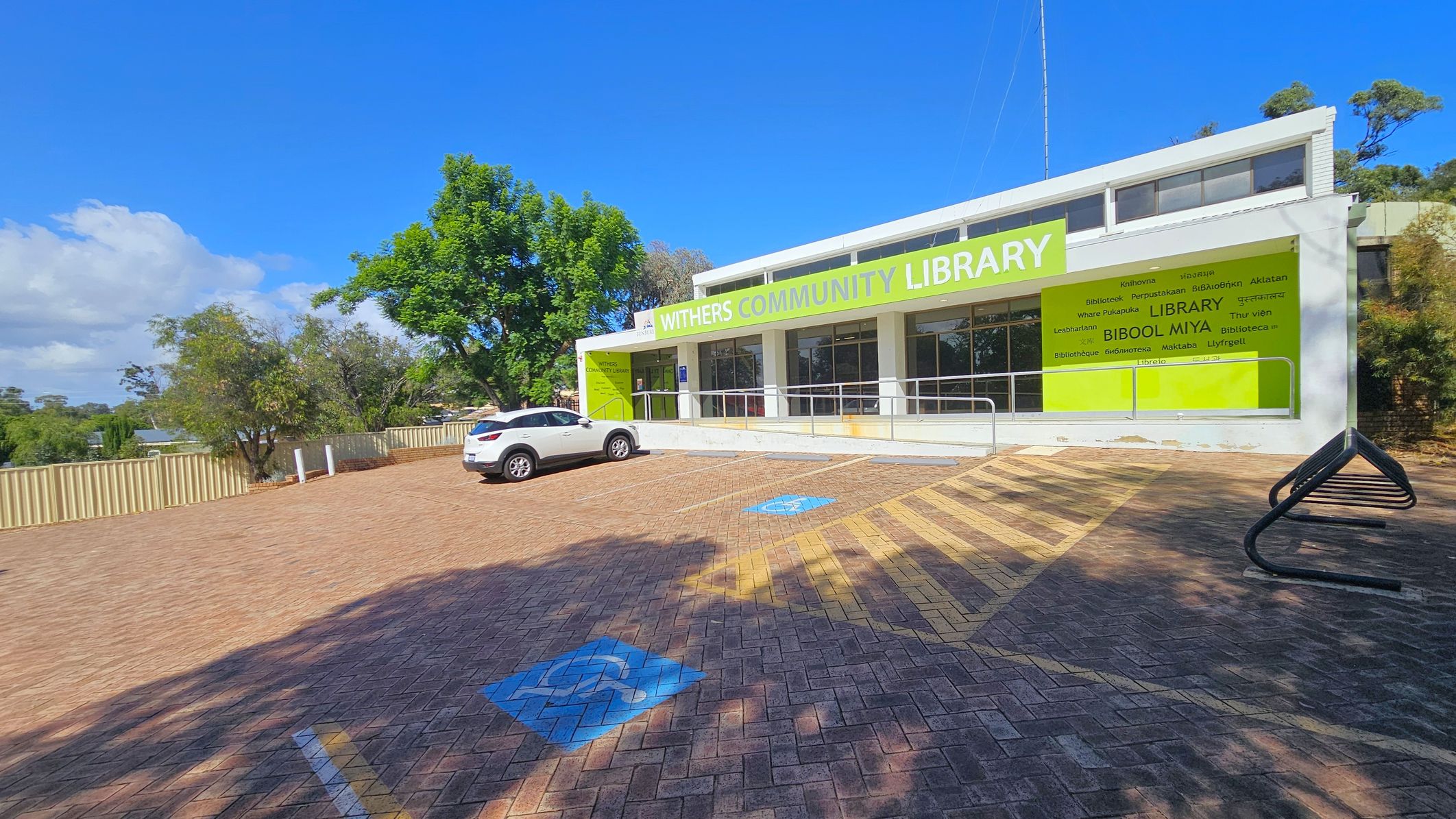 A single-storey building with a large green sign reading "Withers Community Library" on the right. The building has a white facade with multiple windows. In front, there is a paved parking area with a white car parked on the left. Blue disabled parking symbols are visible on the ground. A metal bike rack is on the right. Trees and a beige fence line the background, with a clear blue sky above.