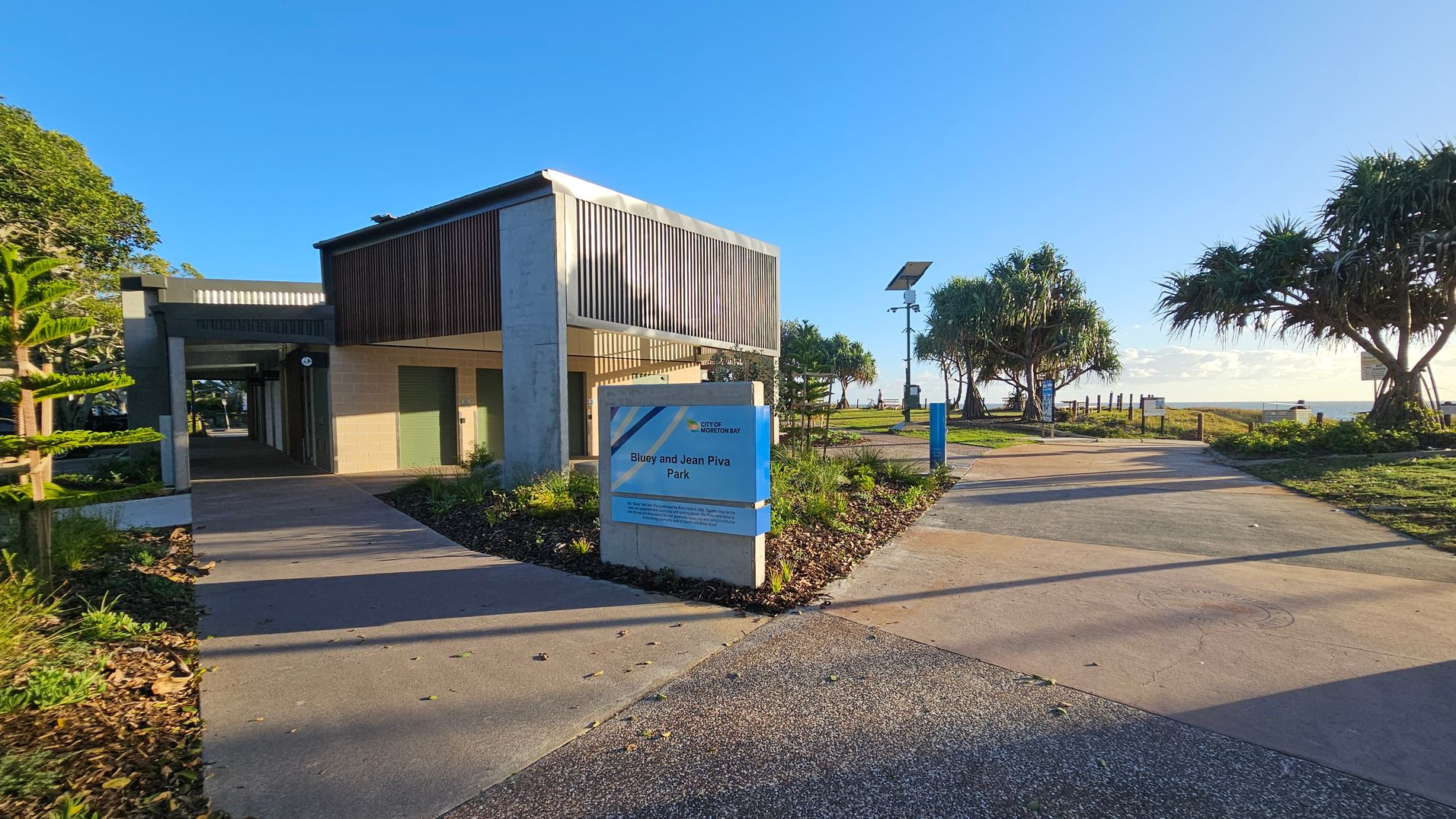 A modern building with vertical slats on the left, featuring a covered walkway. In front, a sign reads "Bluey and Jean Piva Park." The path leads to the right, bordered by well-maintained greenery and palm trees. A solar-powered light pole is visible along the path, which extends towards the horizon. The ground is paved with a mix of concrete and decorative patterns.