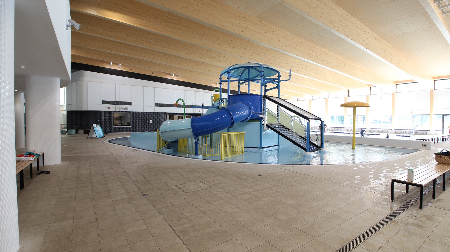 This picture shows the other side of the large aquatic centre. Kids can engage in a variety of activities while at the pool, thanks to the colorful waterpark sliding area and other kid-friendly playthings, with shades of blue and yellow. The pool has light brown lines-like ceiling that matches the multi-coloured splash park. The floor tiles also have a touch of brown. There are available long chairs on the sides of the waterpark.