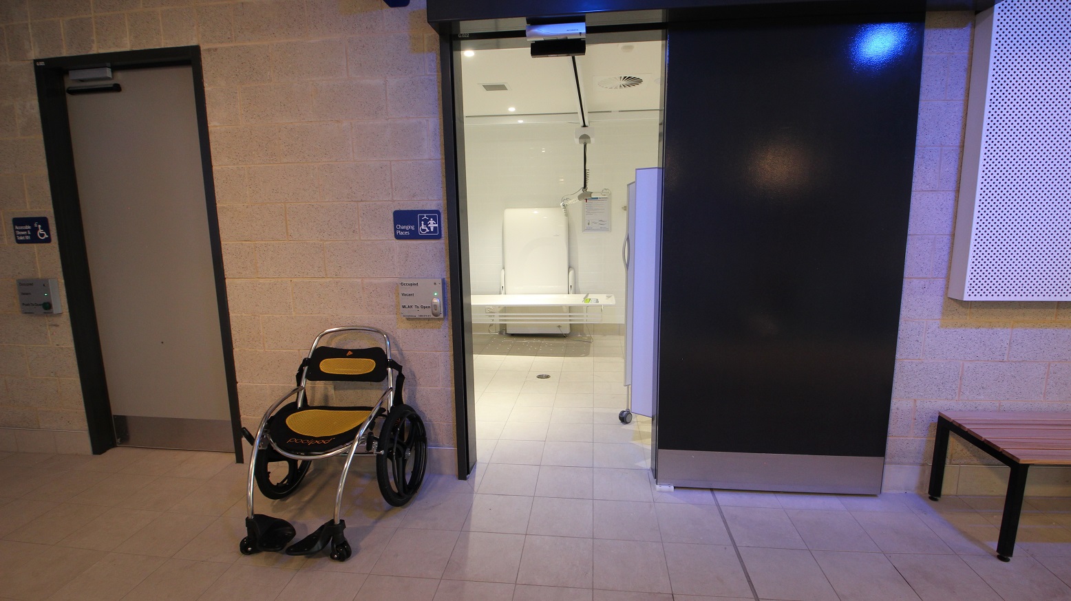 This is a glimpse of what’s inside the changing room that’s all white. What you will see while standing outside the door is the wall support and frame and the toilet bed that can be electrically raised and lowered at different heights. The room’s digital door lock mounted close to the door shows whether it is occupied or vacant and a button to open the room. A wheelchair that has rubberized seat and back support that has a combination of black and yellow color is placed outside.