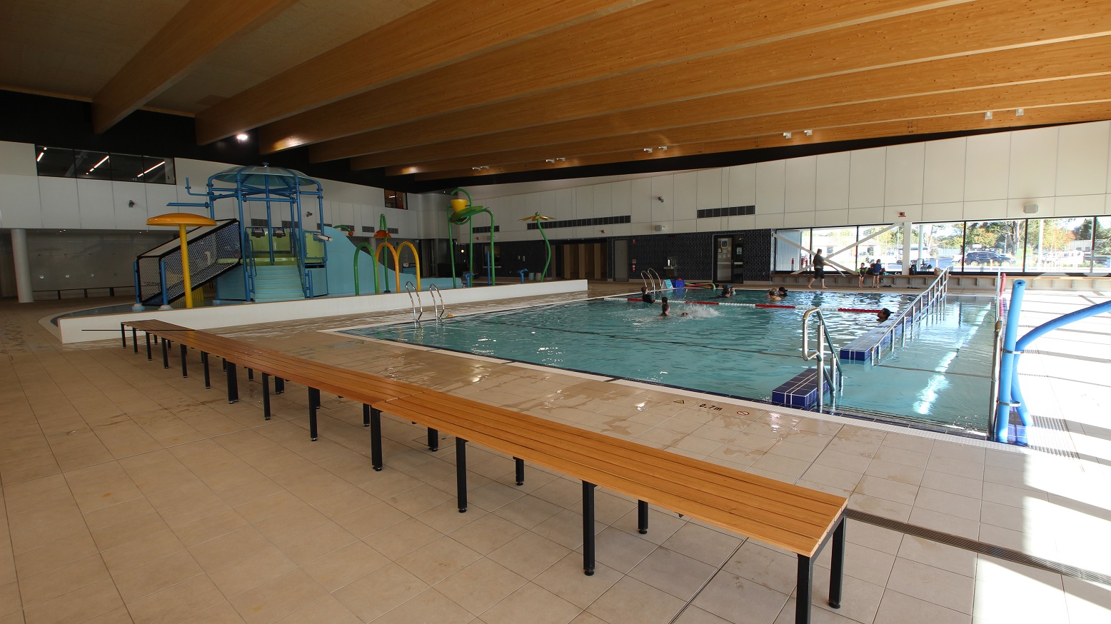 This is the view of the splash park and a swimming pool that extends from one end of the area to the other. Variety of activities for kids can be done at the waterpark pool as it has a kid-friendly sliding area and other playthings available. Three connected long wooden chairs with metal legs are also available near the big pool. Overall, this section of the aquatic centre offers an enjoyable place both for adults and kids.