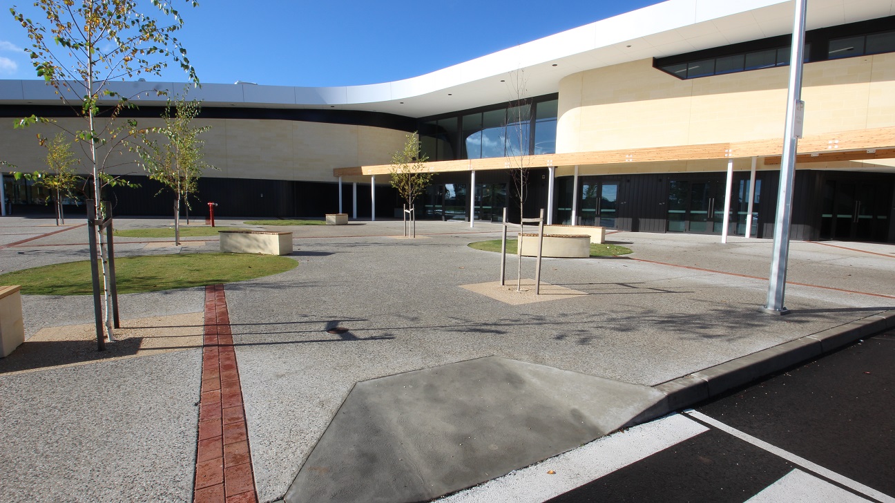 A closer look of the right front view of the building that’s low set, designed with natural colour schemes with creams, whites and grey. Some newly-planted trees and round and rectangular brown benches can be seen on this front view. The trunks of the trees are thin and plant braces are still attached to the lower part of the trees. There are also round grassed landscapes that offer a green view. The outdoor flooring has mostly grey surfaces with red linings.