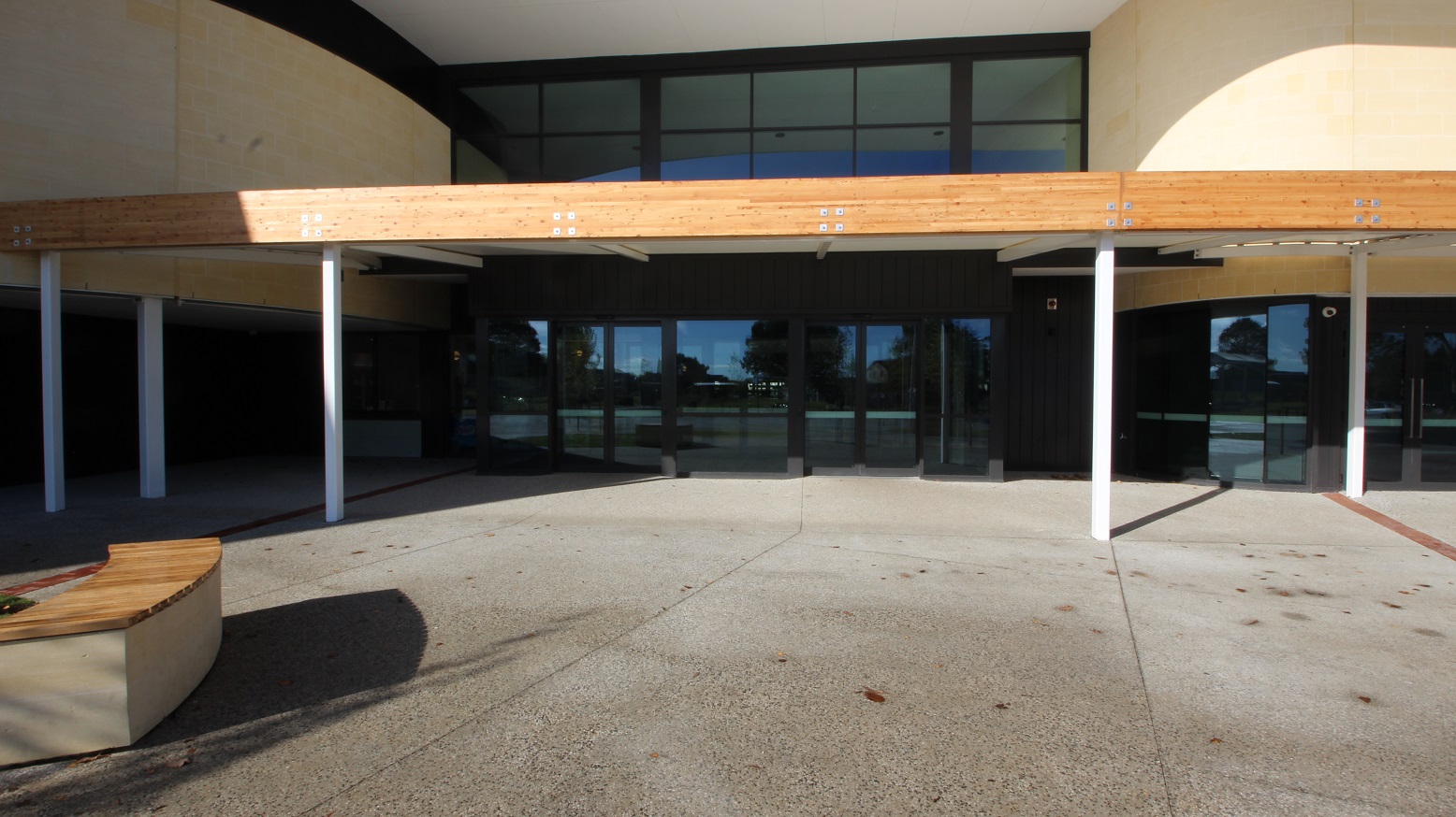 A closer look of the front right view of the building that shows the glass wall and doors and the walkway roof that’s made of wood supported by the white pillars. A nearby bench is shown in this view that has beige and brown surfaces.