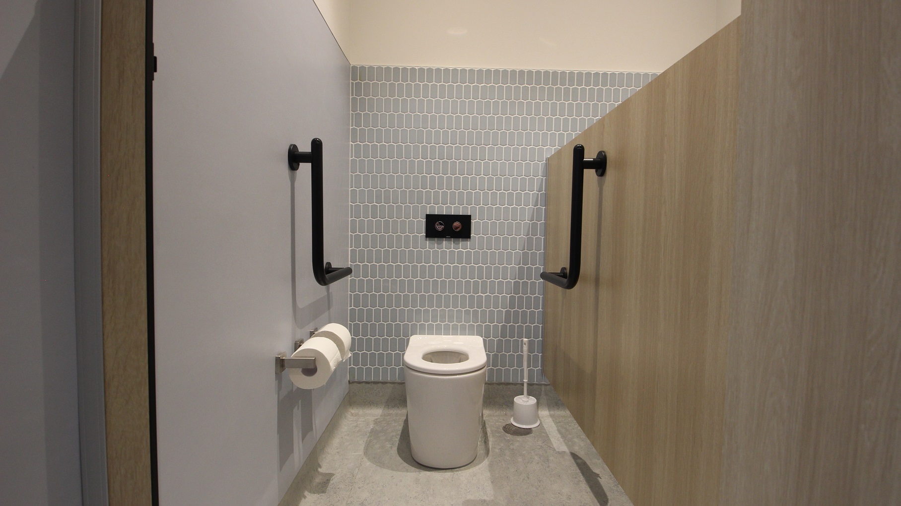 The inside view of the ambulant toilet that showcases three different wall surfaces. The right wall has a plain grey wall where the tissue holders are mounted. The back wall is designed with small grey tiles. It has a white toilet bowl with a black push button for the flush function and black handrails on both sides. The brown wall to the right is actually the partition of the two toilet units. There is also a toilet bowl cleaner placed in the corner.