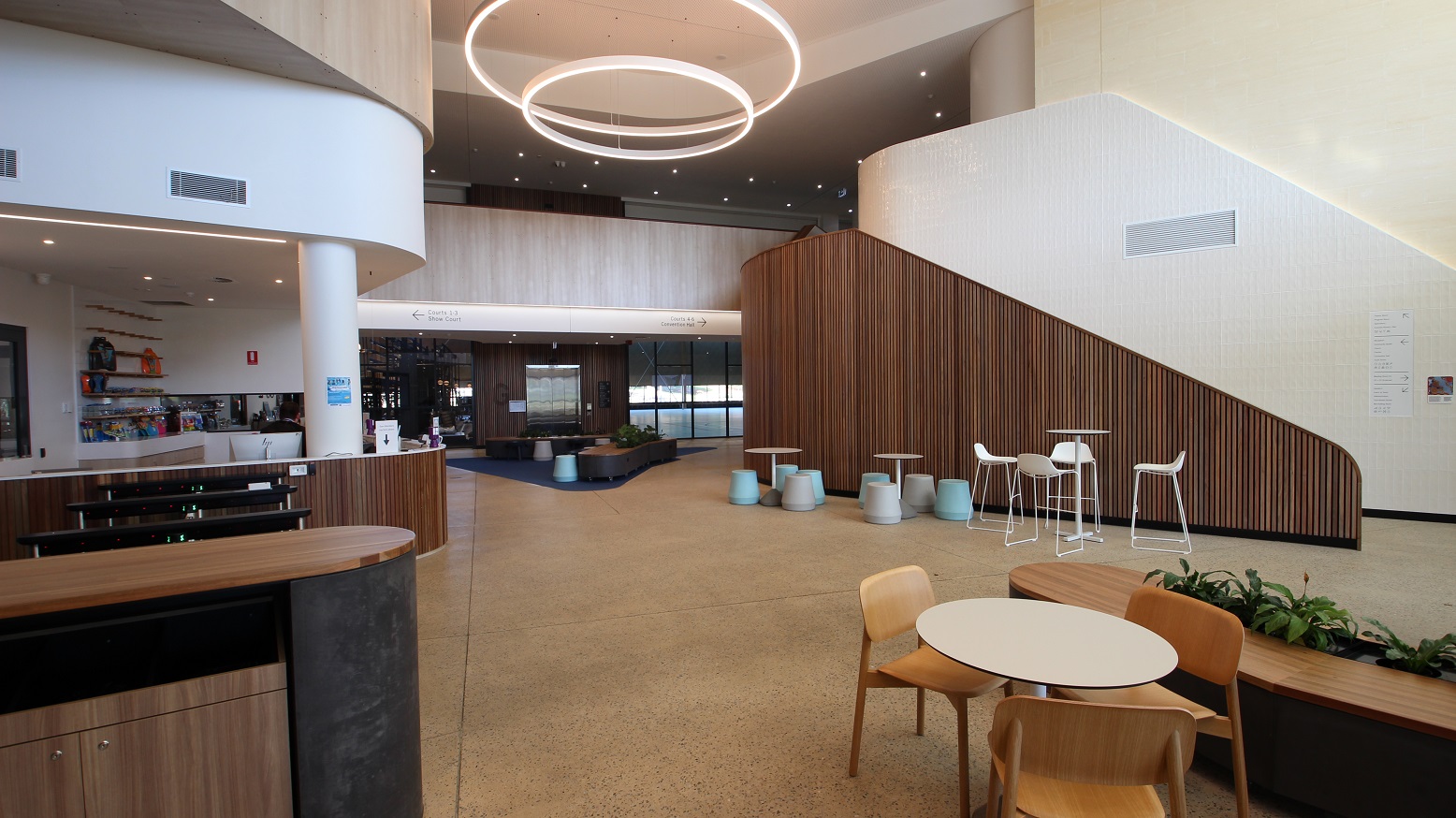 This is the view near the main entrance where you can see the lobby and the hallway going to the other sections of the center. Near the island counter is a set of a white table with white chairs and next to it is the built-in plant shelf of the lobby bench. In the middle of the hallway is a large white circular fluorescent ceiling light above a timber-clad stairwell to the second floor. More tables and chairs are available aligned to the wooden stairwell partition.