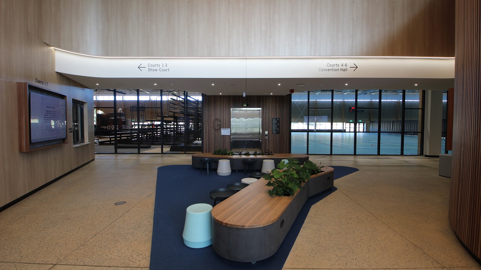 This is the middle section of the hallway where there are two more lobby benches available and white, black and pastel blue-green chairs. Walking straight to the hallway will lead you to the elevator with a wooden design and has glass walls on both sides. It has the big letter G on the left and the right side has the building directory. There is a huge monitor at the left side of the lobby benches with a sign above saying “Courts 1-3”.