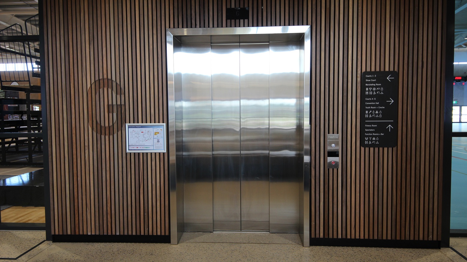 This highlights the modern wood clad cover design of the elevator with silver door. You will be able to see the other sections of the centre through the glass walls on both sides of the elevator. A directory list of rooms and sections of the building is also provided and placed on the right side of the elevator door. There are corresponding arrows pointing to where you should go such as a fitness room, function rooms, bars and a lot more.