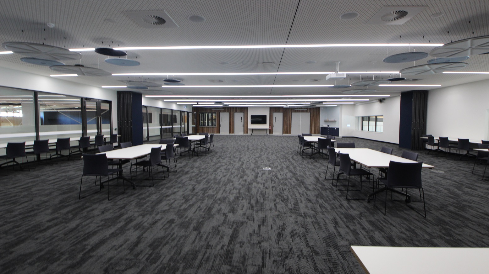 This is the view of function room 1 from its main door. Immediately on the left side of the door is the signage that says “Function Room 01” with a people sign icon on it. The door’s casing is black with silver door handles. The function room is large enough that guests have a wide space good enough to walk around. The room view looks pleasant as the bright fluorescent lighting and white and blue round ceiling ornaments is a contrast to the grey carpeted floor.