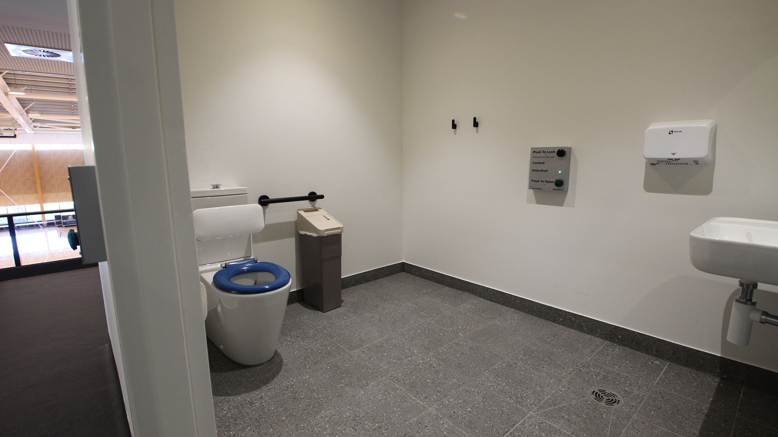 The inside view of the toilet with white walls and grey tiles. Immediate to the left is the white back to wall toilet suite that has a blue seat lid to assist guests with low vision. There is also a black trash bin with a beige cover and above it is the black grab rail. The wider wall of the toilet has two black hooks and a mounted hand dryer. In the middle part of this wall, you will find a grey digital door lock.