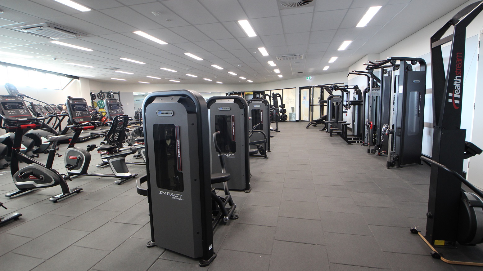 An open glass door that shows the different fitness equipment such as cardio-vascular machines, resistance weights, and more. The dark floor has a good contrast with low white ceilings and bright fluorescent lights. The room’s right door has some papers glued on it such as colourfully plotted gym schedule and timetable. The left side has the fitness room signage with a combination of white and black design.
