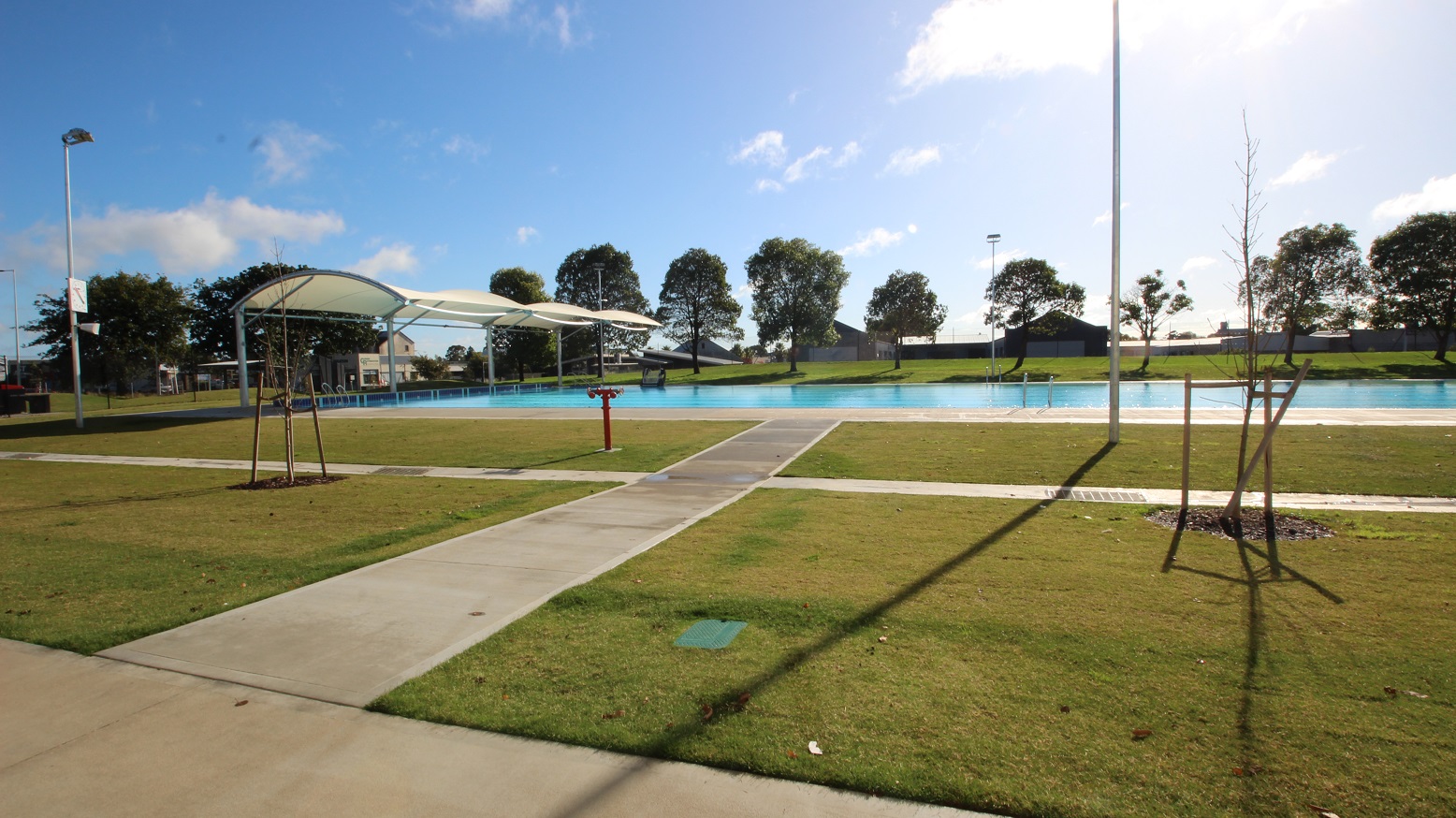 The other side of the outdoor pool reflects eight pool lanes with black markings on the pool floor. The expansive grassed landscaped scenery on the other end of the rectangular pool creates a relaxed and casual setting. The shorter side of the pool has the canopy that provides shade to the guests who stay on this side. The other two sides of the pool have the tiled flooring. From afar, a low-set building can be seen.
