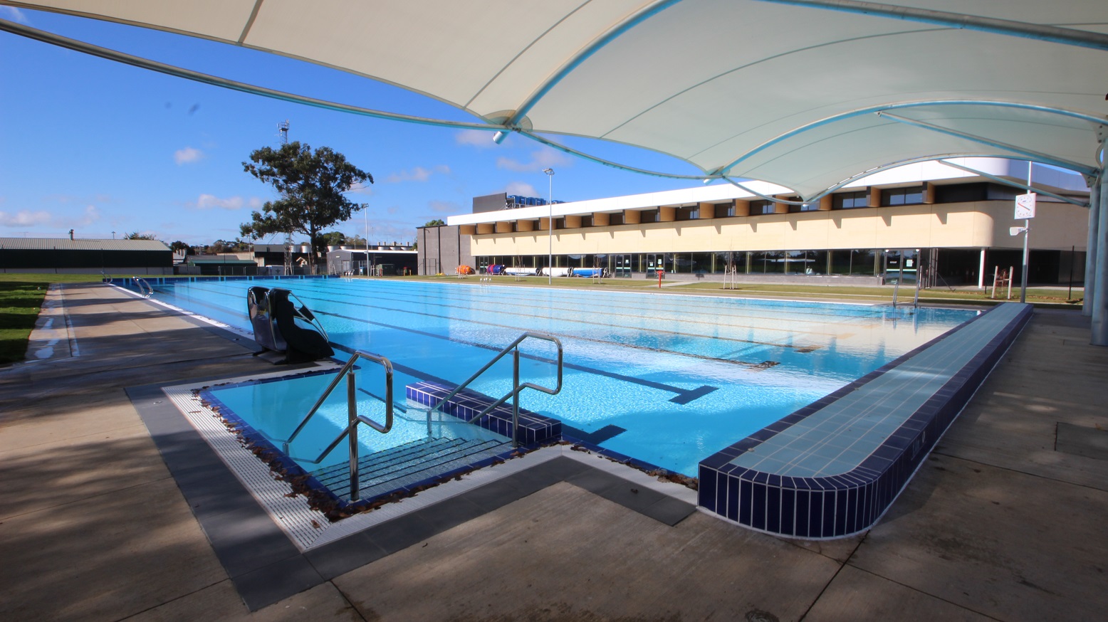 This outdoor pool has an access pool stair on its left, a pool pod and a white canopy that looks like a half tent. The other two sides of the pool have tiled flooring. From a distance, a low-set building can be seen. On the other end of the rectangular pool, the expansive grassed landscaped scenery reflects eight pool lanes with black markings on the pool floor.