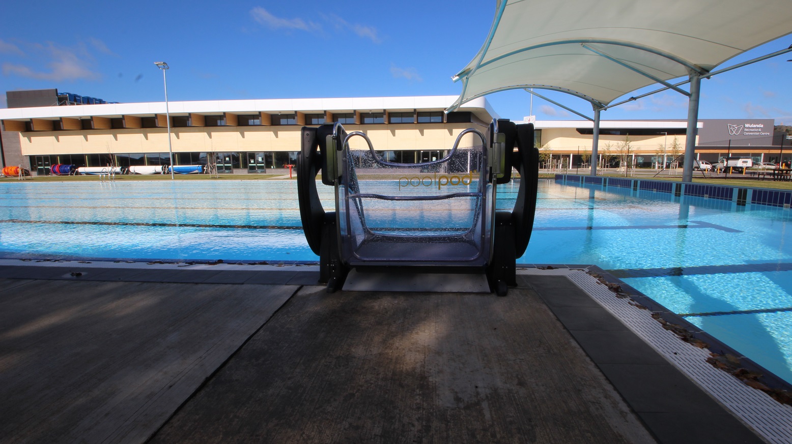 This outdoor pool showcases a pool pod that is placed on the side of the pool access stair. A white canopy is built on the right side of the pool. The lane markers are reflected on the water with long letter T-like black markings. A refreshing view from this side lets you see the indoor aquatic center and the building façade having the blue sky as its background.