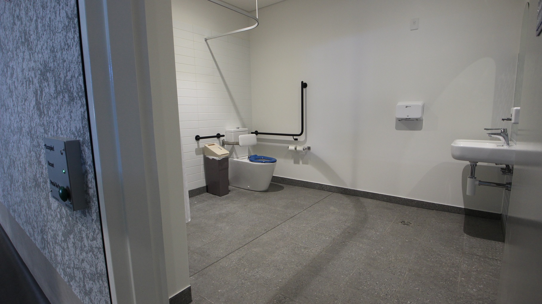 The inside view of the stadium’s toilet with white walls and grey tiles. From the door’s view you will see at the opposite corner the white back to wall toilet suite that has a blue seat lid and a white back support. Near the toilet bowl are inverted L and a straight black grab handles and tissue paper and its holder mounted on the wall. There is also an extendable corner curved shower rod with white curtain, a white sink with liquid soap dispenser.