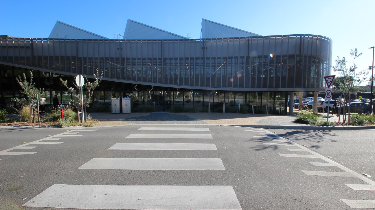 Zebra crossing leading to Yawa Aquatic Centre. Plant in a plant box on the right side near the fully packed parking space located on the rear end of the facility is a 10-kilometer per hour speed limit and a roundabout traffic sign. Several curvy metal bike racks and perforated metal bin enclosures with a Mornington Peninsula logo are available within the pathway that surrounds the facility.