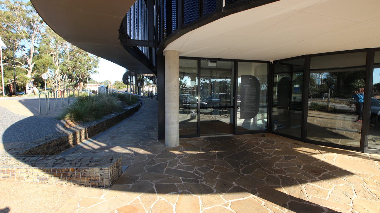 Near the curvy metal bike racks and the perforated metal bin enclosures at the rear end of the facility is a railed staircase leading to the river stone floor design that connects to the automatic, double glass entry doors with black metal edges. Found on the glass of the doors are the contact information and opening hours and a black poster about the facility, while a CCTV camera can be found mounted on top of the glass doors.